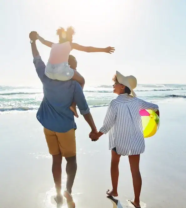 Family walking on beach. The woman is holding the man's hand, looking at him, while he carries the child on his shoulders.